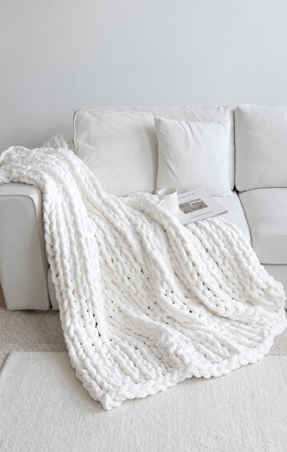 A complete white chunky knitted blanket draped over a white sofa with a mug and magazine